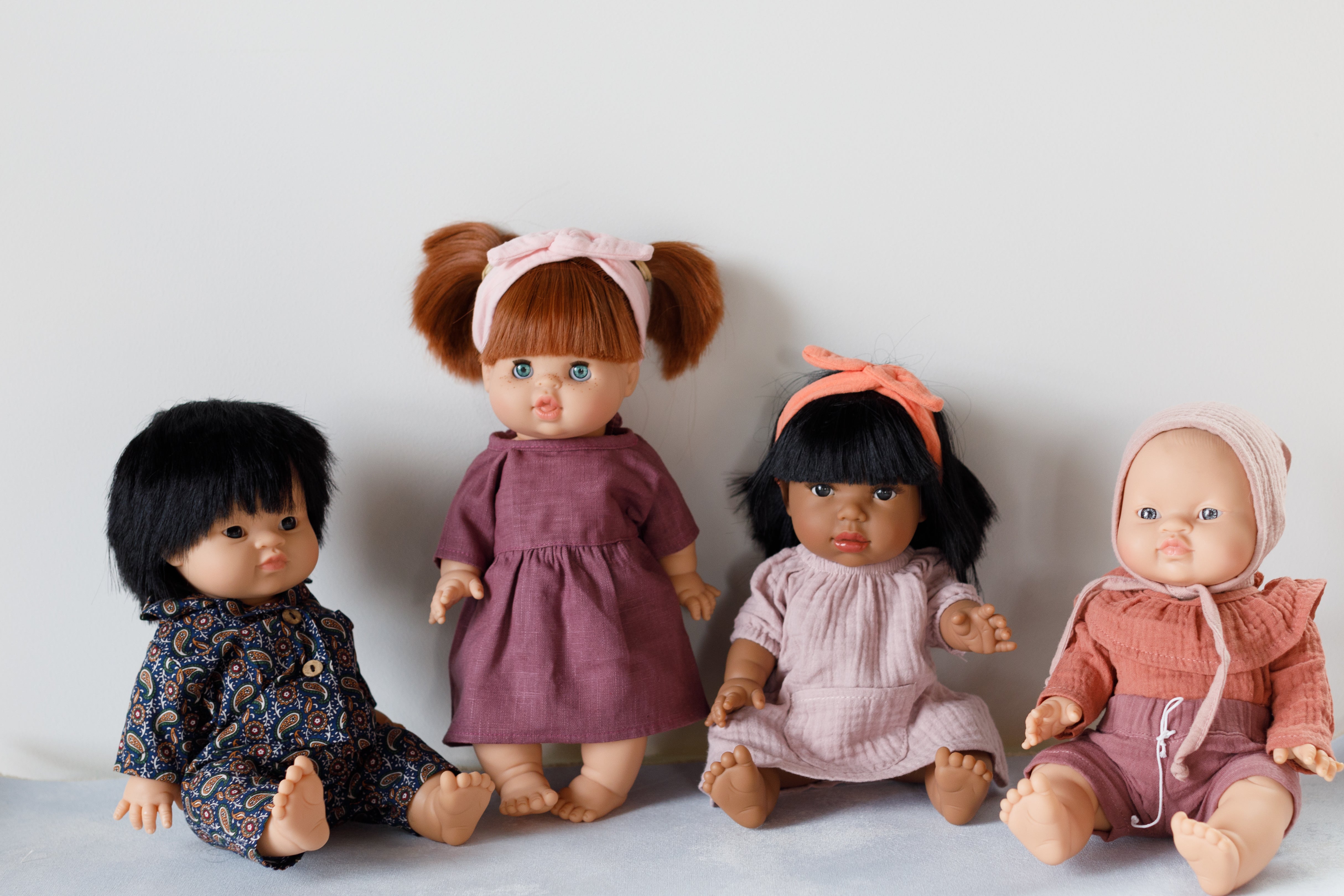 Four dolls are sitting in a row. Each doll has a different outfit and hairstyle. The background is plain, and the surface they sit on is light-colored.