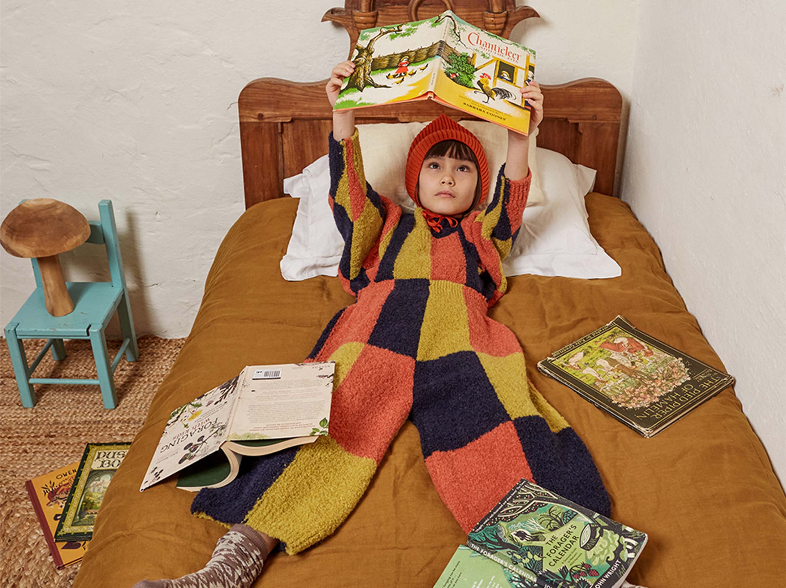 Child in colorful checkered outfit and orange hat lies on a bed, holding a book overhead. Surrounding them are more books, a small blue chair, and a mushroom-shaped lamp.