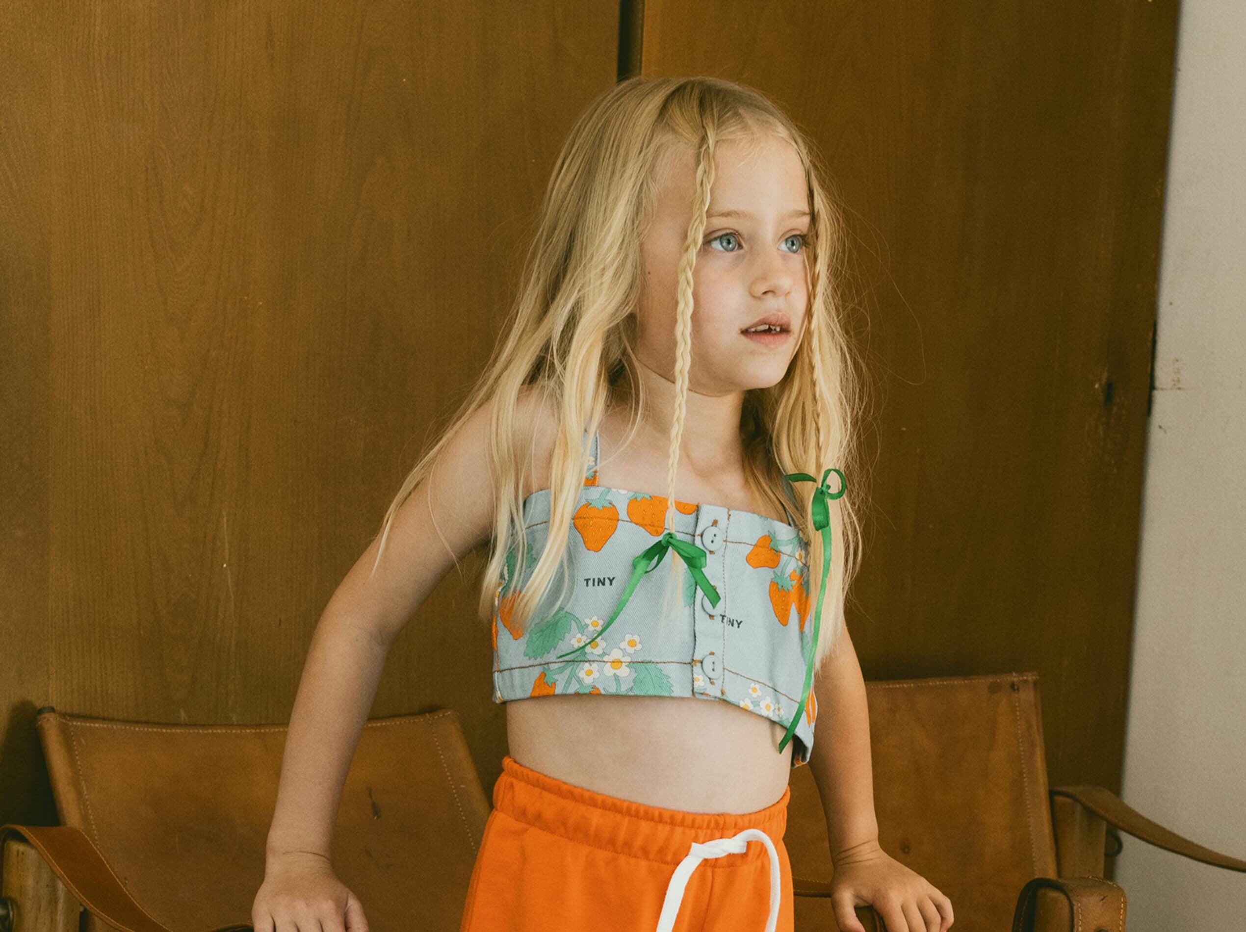 Child with long blonde hair stands in front of a wooden background, wearing an orange skirt and a green top with orange patterns.