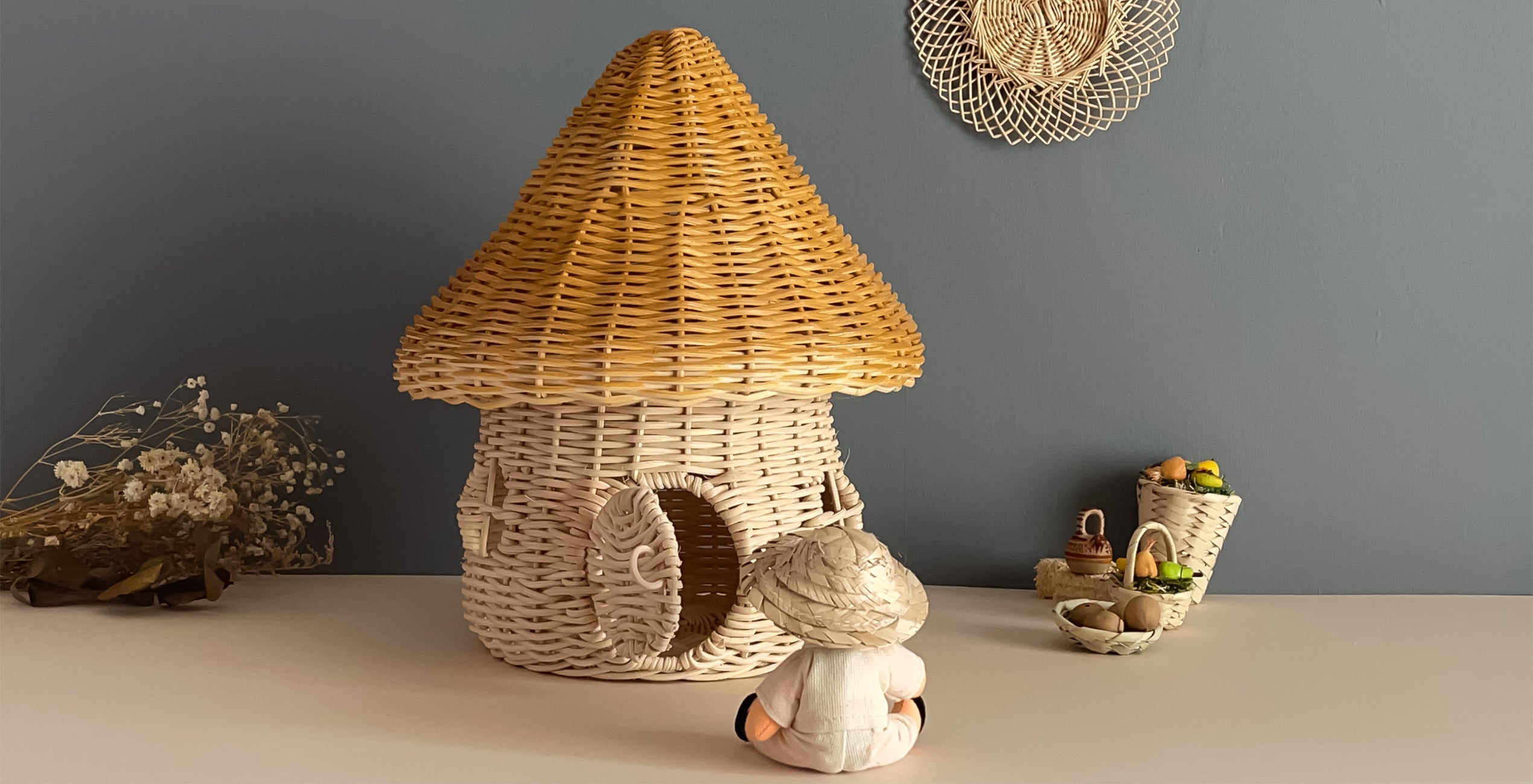 A small wicker house with a conical roof is placed on a table. A white figurine sits before it. In the background are decorative items including dried flowers and a basket filled with small objects.