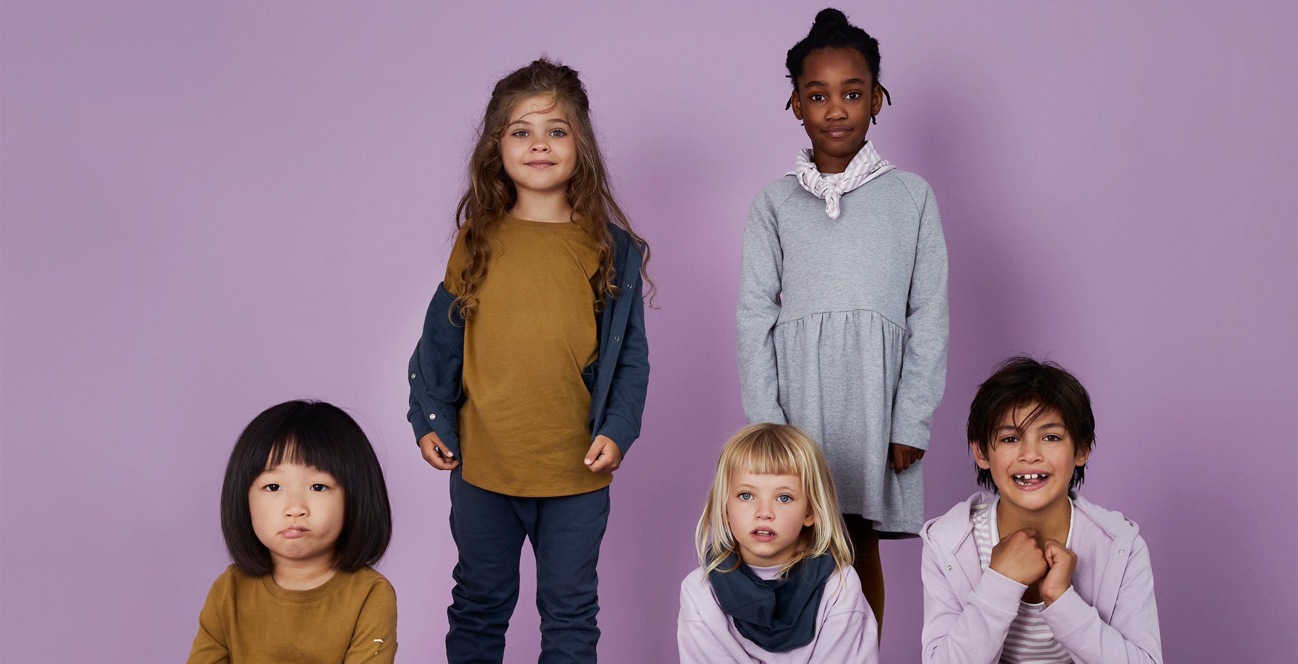 A group of five children stand and sit against a plain purple background. They are dressed in casual clothing in various shades of yellow, gray, and purple.