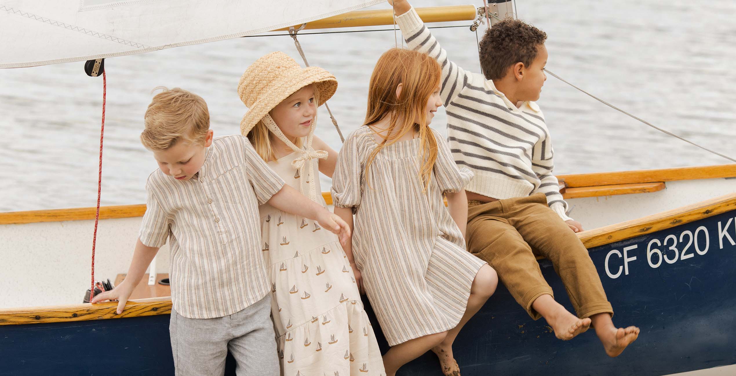 Four children, dressed in neutral-toned clothing, sit on the edge of a boat with a white sail on a body of water. One child wears a straw hat, and the boat's registration number "CF 6320 K" is visible.