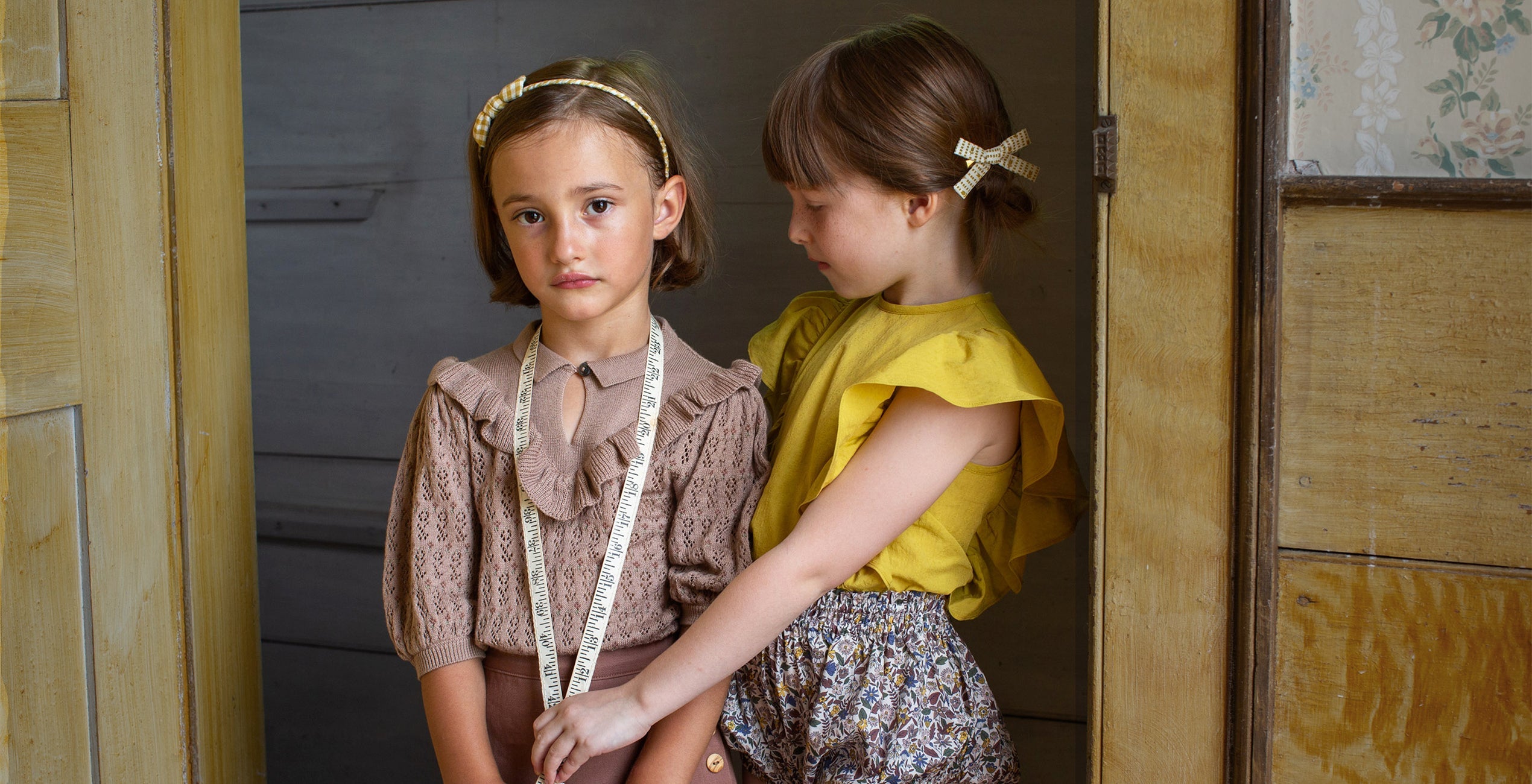 Two young girls stand indoors by a wooden door, one wearing a headband with a measuring tape around her neck and the other in a yellow top tying the tape's end.