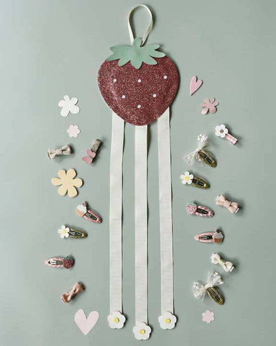 The sweet strawberry 3 ribbon clip hanger by Rockahula Kids features a decorative strawberry shape with whimsical flower and heart cutouts, hair clips, and bows on a light green background, offering a perfect blend of charm and style!.