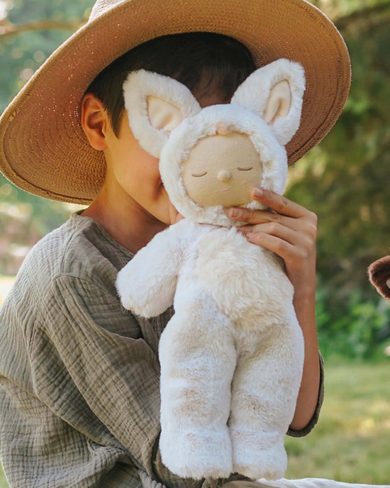 A child wearing a wide-brimmed hat holds their Olli Ella Cozy Dinkum Fox Fenix plush toy, seated outside with greenery in the background.