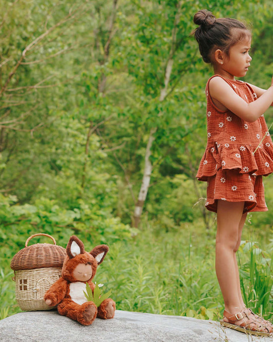 A young girl in a red dress stands on a rock, holding a stick. Next to her are an Olli Ella Cozy Dinkum Fox Finnley plush toy and a wicker picnic basket, surrounded by lush green foliage that creates an enchanting scene.