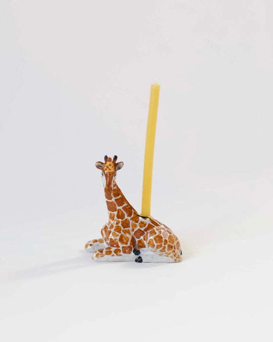 A hand-painted ceramic camp hollow giraffe cake topper with a yellow candle positioned on its back, set against a plain white background.