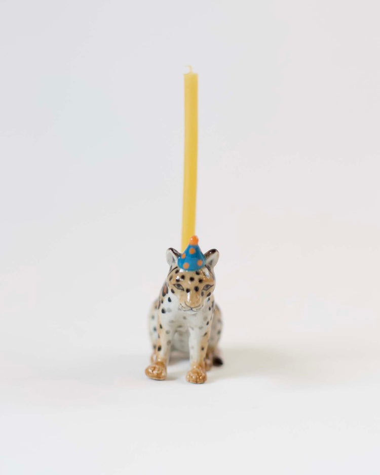 A small figurine of a Camp Hollow jaguar cake topper with a yellow candle balanced on its head, against a plain white background.