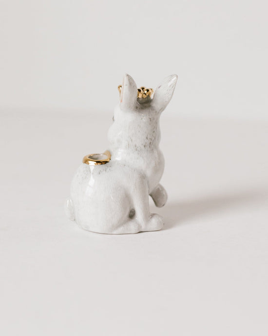 A hand-painted ceramic Royal White Rabbit cake topper with two gold rings placed on its ears, set against a plain white background by Camp Hollow.