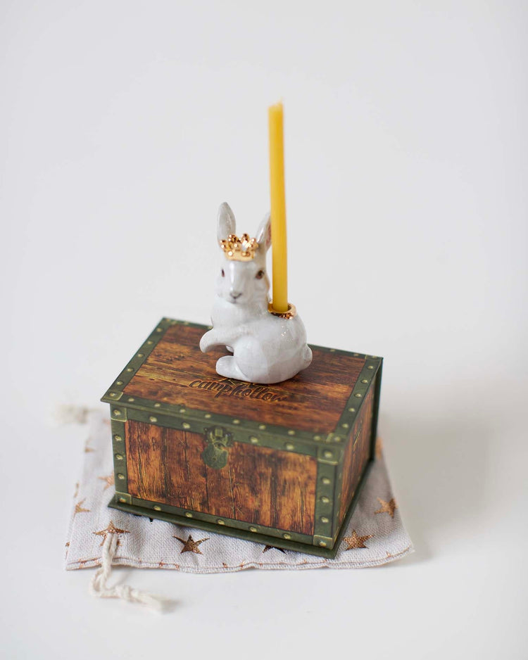 A decorative candle holder shaped like a Camp Hollow Royal White Rabbit Cake Topper sitting atop a vintage wooden box, with a lit yellow candle.