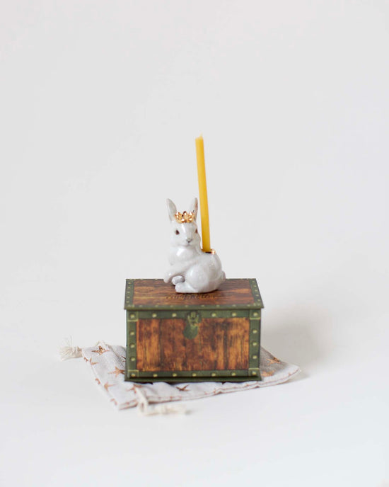 A small ceramic figurine of a royal white rabbit cake topper, hand-painted and perched on a decorative box with a yellow candle on its back, set against a plain white background by Camp Hollow.