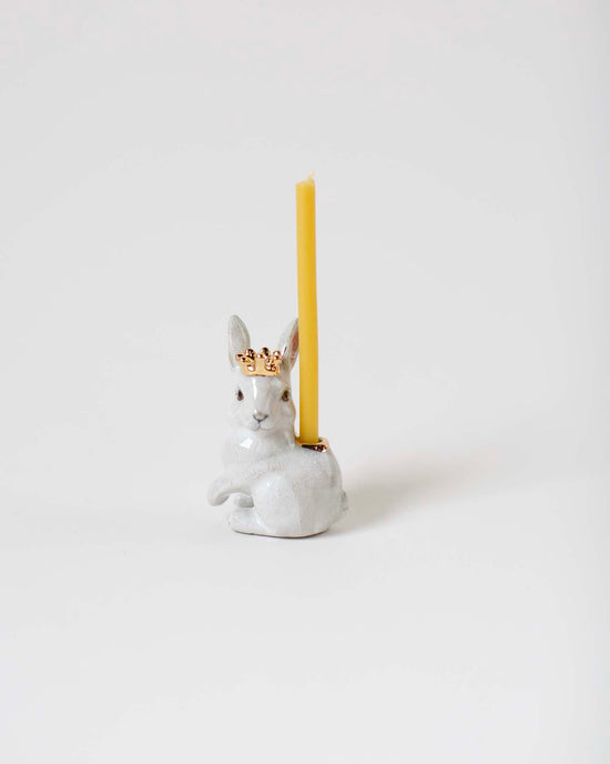 A royal white rabbit cake topper from Camp Hollow, shaped like a white rabbit wearing a golden crown, hand-painted with heirloom-quality craftsmanship, with a yellow candle inserted on top, against a plain white background.