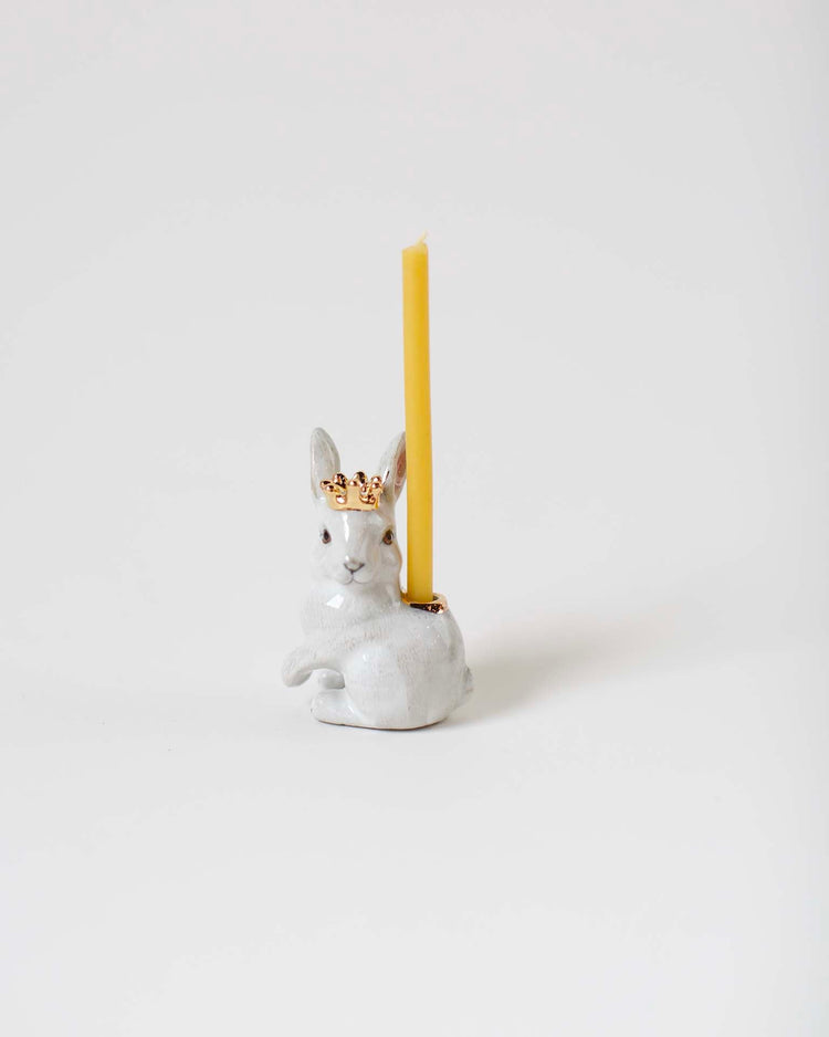 A royal white rabbit cake topper from Camp Hollow, shaped like a white rabbit wearing a golden crown, hand-painted with heirloom-quality craftsmanship, with a yellow candle inserted on top, against a plain white background.