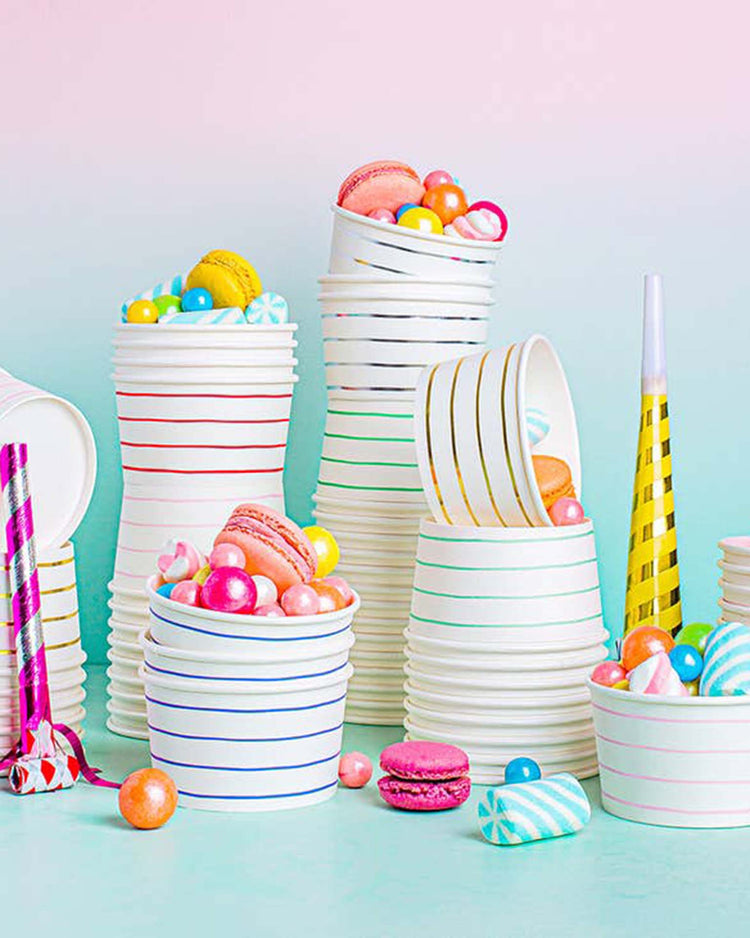 Stacks of Daydream Society's Gold Frenchie striped treat cups overflowing with colorful macarons, candy, and party blowers sit against a pastel background.