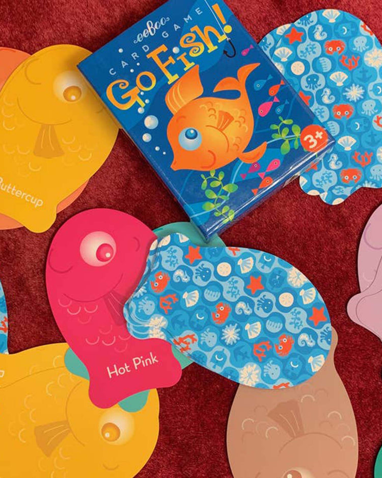 The "color go fish playing cards" by eeboo are displayed on a red surface. Surrounding the central game box are multiple fish-shaped cards in various colors and patterns, making this children's card game an ideal first choice that combines fun with color matching skills.