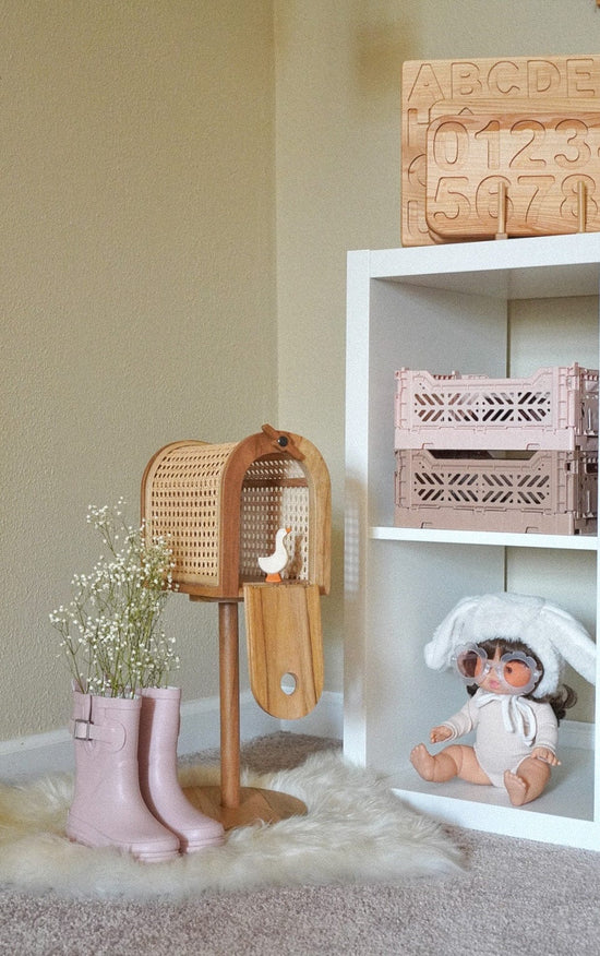 A wooden toy birdhouse, a Poppie Mailbox - Out of Stock ETA OCT by poppie toys for early writing and reading, pink rain boots with white flowers, a doll in a bunny hat, and wooden blocks with letters and numbers are displayed in a cozy, light-colored room.